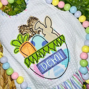May include: A white waffle-textured garment featuring an Easter-themed applique of a basket filled with a gold glitter butterfly, a tan bunny silhouette, a carrot, and blue eggs. The basket has a green grass trim and a blue label reading Demi.