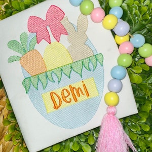 May include: A white fabric square featuring an embroidered light blue egg shape containing a carrot, a yellow egg, a tan bunny, and a red bow. The name Demi is embroidered in orange on a yellow rectangle. A string of pastel wooden beads with a pink tassel.