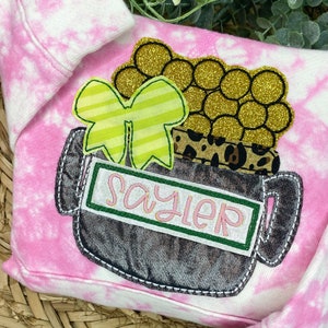 May include: A pink and white tie-dye sweatshirt featuring an appliqué of a dark grey pot filled with gold glitter circles. The design includes a lime green striped bow, a leopard print band, and a white rectangular patch with the name Sayler.