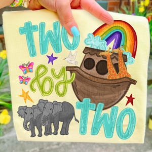 Può includere: T-shirt giallo chiaro con la scritta "TWO by TWO" in turchese e verde. Il design include un arcobaleno, un'arca con giraffe, farfalle, elefanti e stelle.