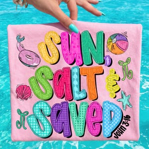 May include: Pink t-shirt with the words "SUN SALT SAVED" in colorful, bubble letters. The design includes a beach ball, starfish, seashell, and a pink inflatable tube. The text "John 3:16" is also visible.