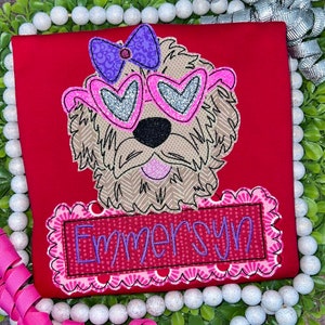 May include: A red t-shirt featuring an embroidered applique of a tan dog wearing pink heart-shaped glasses and a purple bow. Below the dog, a rectangular patch with a scalloped pink border displays the name Emmersyn in purple script.