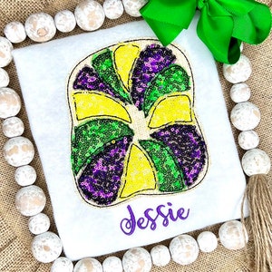 May include: A white t-shirt featuring a large, segmented king cake applique decorated with yellow, green, and purple sequins. The name Jessie is embroidered in purple script below the applique, and a bright green ribbon bow is attached at the top.