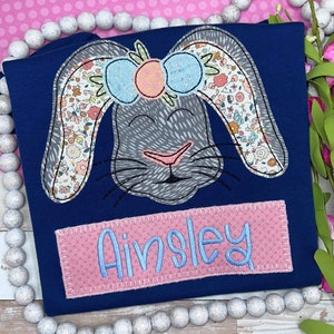 May include: A navy blue t-shirt featuring an embroidered grey rabbit face with long ears decorated in a floral pattern and a light blue bow. Below the rabbit, a rectangular pink patch is stitched onto the fabric with the name Ainsley in blue.