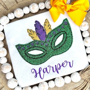 Op de afbeelding: Een wit t-shirt met een groene glitterende maskerapplicatie met paarse en gouden veren. De naam Harper is in paars cursief schrift onder het masker geborduurd. Een felgele strik is aan de bovenhoek van het t-shirt bevestigd.