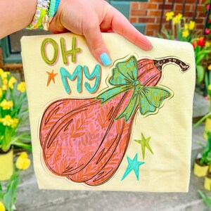 May include: A pale yellow t-shirt featuring a graphic of a pumpkin with a pink and orange leaf pattern, adorned with a large teal bow. The text "OH MY" is printed in teal and green above the pumpkin, accompanied by small teal and green stars.