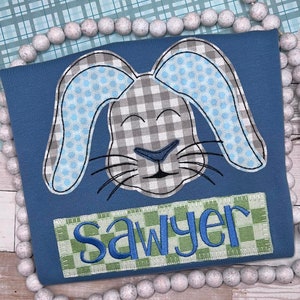 May include: A blue t-shirt featuring an embroidered rabbit head with grey and white checkered patterns and light blue honeycomb accents. Below the rabbit, a rectangular patch displays the name Sawyer in blue script on a green checkered background.