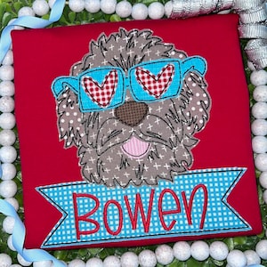 Op de afbeelding: Een rood t-shirt met een applicatie van een bruine hond met een wit kruispatroon, die een felblauwe bril met rood-witte hartvormige glazen draagt. Onder de hond staat een blauw-wit geblokte banner met de naam Bowen in rode letters.