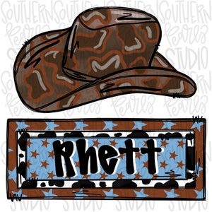 May include: A brown cowboy hat with a dark brown and tan cow print pattern positioned above a rectangular sign featuring the name Rhett in black letters, set against a light blue background with brown stars and a black and white cow print border.