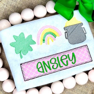 May include: A white St. Patrick's Day themed applique with a green shamrock, rainbow, and pot of gold design. The name "ANSLEY" is embroidered in green on a pink and white plaid patch. A green bow is at the top.