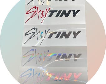 May include: Five white stickers with the text "Stray Kids" in a stylized font and "Tiny" in a bold font. The stickers are arranged vertically, with the top sticker featuring black text, the second sticker featuring red text, the third sticker featuring silver text, the fourth sticker featuring holographic silver text, and the bottom sticker featuring holographic rainbow text.