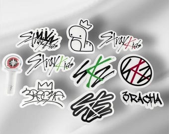 May include: A collection of white stickers with black and green text and graphics. The stickers feature the words "Stray Kids" and other related designs, including a cartoon character with a crown and a light stick.