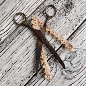 May include: A pair of rusty scissors with a brown crocheted lace ribbon on a white wooden surface.
