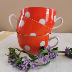 May include: Three orange ceramic mugs with white polka dots stacked on top of each other. The mugs have a white rim and a white handle. The mugs are sitting on a white surface with a bouquet of purple flowers.