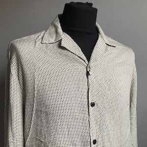 Puede incluir: Una camisa blanca de manga larga con un patrón geométrico negro. La camisa tiene cuello y cierre de botones en la parte delantera.