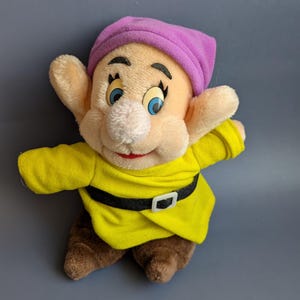 Op de afbeelding: Een knuffel van het personage Dopey uit Sneeuwwitje, voorzien van een lichtpaarse muts, een felgele tuniek met een zwarte riem en witte gesp, en bruine schoenen. Het figuur heeft grote oren, blauwe ogen en een zacht, lichtgekleurd gezicht.
