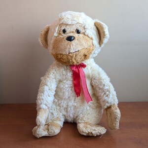 Pode incluir: Um macaco de pelúcia com pelo creme, rosto, orelhas e mãos castanhas e uma fita vermelha amarrada ao pescoço. O macaco tem olhos pretos e uma expressão sorridente. O brinquedo está sobre uma superfície de madeira.