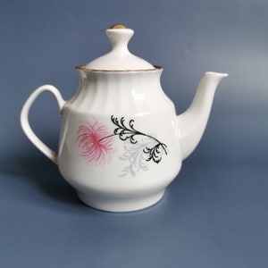 Puede incluir: Tetera de porcelana blanca con tapa y asa curva. La tetera presenta un diseño floral rosa y negro y ribetes dorados. La tapa tiene un adorno dorado. La tetera está diseñada para servir té.