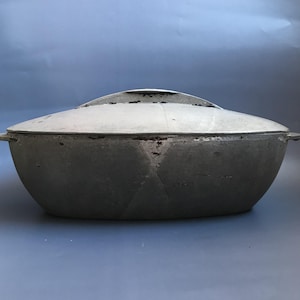 以下が含まれることがあります： 蓋付きの大きな楕円形の鋳鉄製の鍋。鍋はグレーで、わずかにざらざらした表面をしています。蓋の上には小さな取っ手があります。