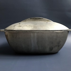 May include: A vintage aluminum oval Dutch oven with a lid. The oven has a slightly textured surface and is a light gray color.