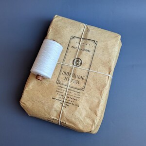 May include: A package of sewing thread wrapped in brown paper, tied with white string, and a spool of white thread. The package has text in Russian. The background is a solid blue-gray.