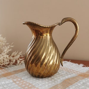 May include: A gold-colored metal pitcher with a swirling design. The pitcher has a wide mouth and a handle.