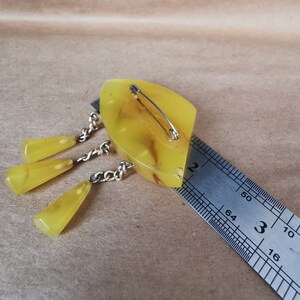 Op de afbeelding: Een broche van geel hars met een hangende ketting en drie gele harsdruppels. De broche wordt gemeten met een liniaal en is ongeveer 5 cm lang.