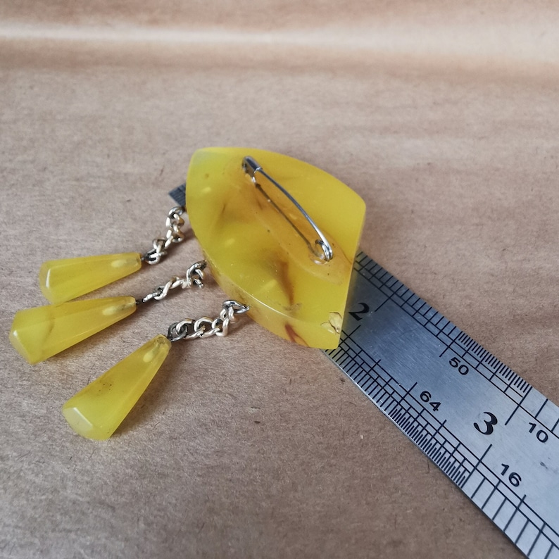 Op de afbeelding: Een broche van geel hars met een hangende ketting en drie gele harsdruppels. De broche wordt gemeten met een liniaal en is ongeveer 5 cm lang.