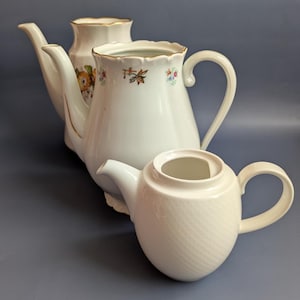 May include: Three white ceramic teapots of varying sizes. The largest teapot has a floral design and gold trim. The middle teapot has a floral design and gold trim. The smallest teapot has a textured, diagonal pattern.