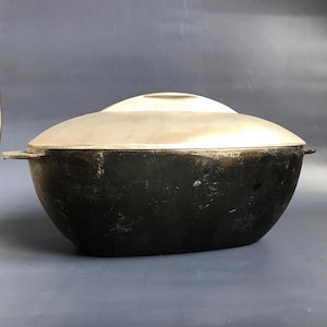 以下が含まれることがあります： 銀色の蓋が付いた黒い鋳鉄製のダッチオーブン。オーブンは楕円形で、両側にハンドルが2つ付いています。