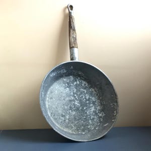 Puede incluir: Una sartén de aluminio vintage con mango de madera. La sartén tiene una superficie gris desgastada con un aspecto moteado.