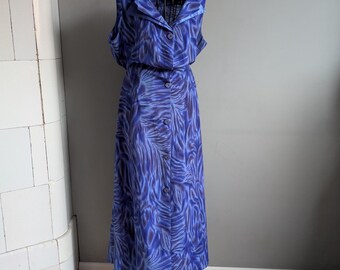Può includere: Un abito blu senza maniche con bottoni sul davanti e colletto. L'abito presenta un motivo di linee astratte blu e viola. L'abito è esposto su un manichino nero.