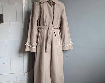 Può includere: Un lungo trench beige con colletto a punta, chiusura frontale con bottoni e cintura in vita coordinata da annodare. Le maniche presentano polsini ampi rifiniti con profili bianchi e un singolo bottone, creando una silhouette strutturata a figura intera.