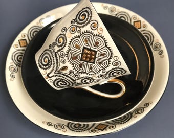 May include: A white teacup and saucer set with black and gold geometric designs. The cup and saucer have a black rim with gold accents.