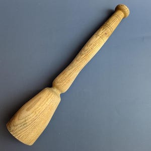 May include: A light-colored wooden muddler with a rounded handle and a flared base. The handle has three carved rings near the top. The muddler is designed for mixing ingredients in cocktails or other beverages.