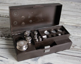 Puede incluir: Una caja de plástico marrón con un juego de pesas de metal plateado para una balanza. La caja está abierta y las pesas están dispuestas en filas.