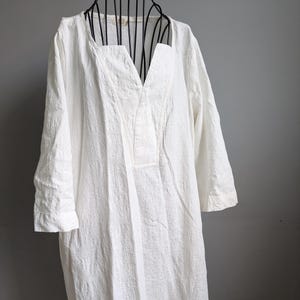 May include: A white, long-sleeved dress with a V-neckline. The dress has a textured fabric and a relaxed fit. The garment is displayed on a black mannequin against a gray background. The dress appears to be made of a lightweight material.