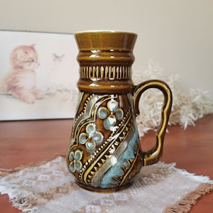 May include: A brown ceramic pitcher with a blue and brown floral design. The pitcher has a handle and a wide mouth.