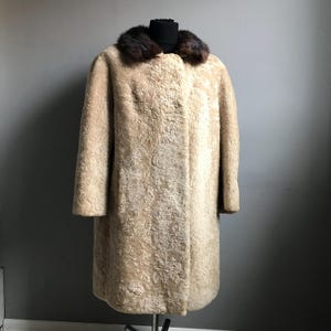 Op de afbeelding: Een lange, cr&egrave;mekleurige schapenvachtjas met een bruine bontkraag.