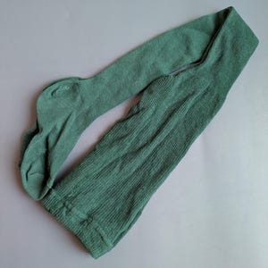 Può includere: Collant verdi, stesi su una superficie grigia. I collant sono di colore pieno, con una trama a coste sulle gambe e una trama liscia sul piede. Il tessuto sembra essere un morbido tessuto a maglia.