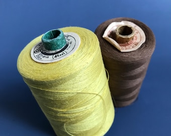 Puede incluir: Dos carretes de hilo, uno amarillo y otro marrón. El carrete amarillo tiene una etiqueta que dice "44/1 250m".