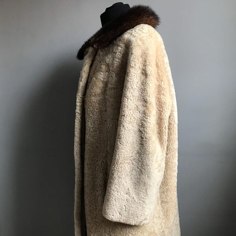 Op de afbeelding: Een lange, cr&egrave;mekleurige schapenvachtjas met een bruine bontkraag. De jas is gemaakt van zacht, pluche bont en heeft een klassiek, tijdloos ontwerp.