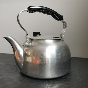 Könnte beinhalten: Ein silberner Aluminium-Wasserkocher mit einem schwarzen Kunststoffgriff und -deckel. Der Wasserkocher hat einen abgerundeten Körper und einen Ausguss an der Seite.