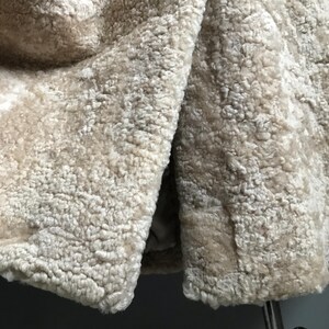 Op de afbeelding: Close-up van een beige nep bontstof met een zachte, pluche textuur. De stof is dubbelgevouwen, waardoor de binnen- en buitenkant van het materiaal zichtbaar is.