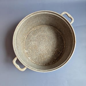 Può includere: Vista dall'alto di una grande pentola rotonda in ghisa con due manici. La pentola è di un grigio opaco e sembra ben usata. L'interno della pentola è visibile, mostrando una superficie strutturata. La pentola è su una superficie grigia.