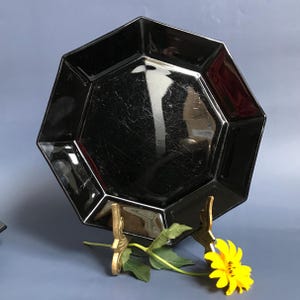 May include: A black octagonal glass plate on a gold metal stand. The plate is on a blue surface with a yellow flower in the foreground.