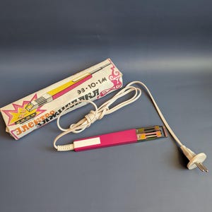 Könnte beinhalten: Ein pinkfarbenes und silberfarbenes elektrisches Lockenstab-Set mit weißem Kabel und Stecker, neben der Originalverpackung. Die Box ist weiß mit rosa und gelben Akzenten und kyrillischem Text.