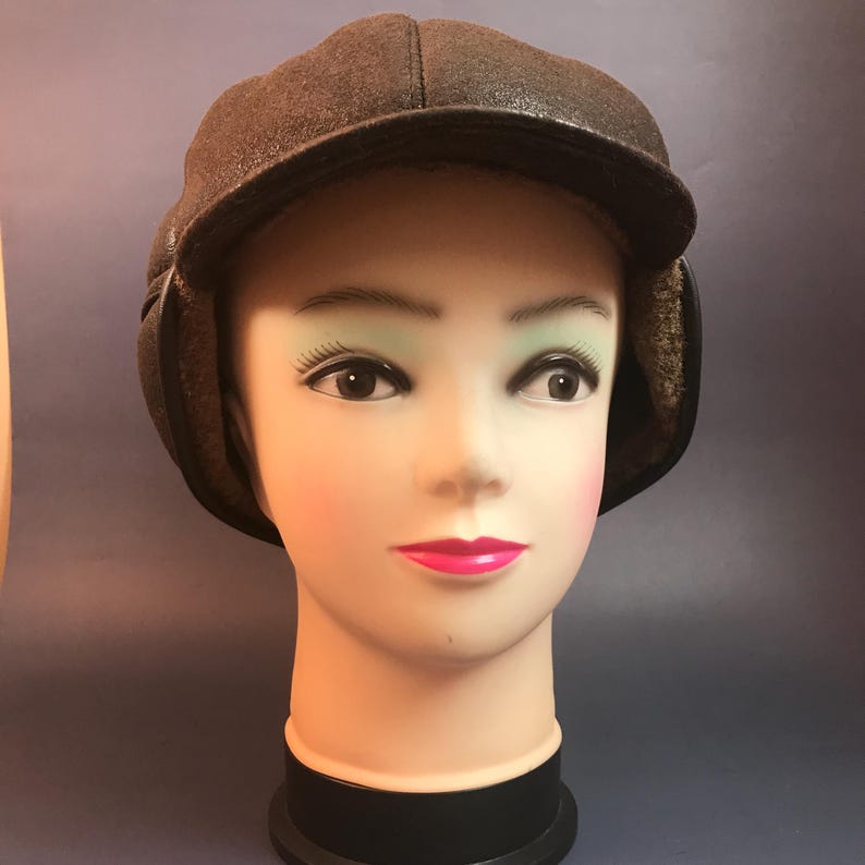 May include: A brown, textured hat with ear flaps, displayed on a mannequin head. The hat has a short brim and a soft, furry lining visible around the ears. The hat is designed to provide warmth and protection from the cold.