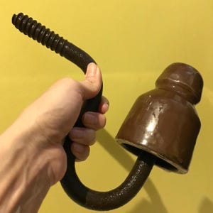 Può includere: Un isolatore in ceramica marrone con un gancio metallico attaccato alla parte inferiore. Il gancio è piegato a forma di U e ha un'estremità filettata.