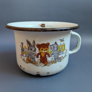 Op de afbeelding: Witte emaille mok met een bruine rand en handvat. De mok heeft een kleurrijke illustratie van een konijn, beer en kat die in een bloemenveld lopen, met een trein op de achtergrond. De mok heeft wat roestvlekken.